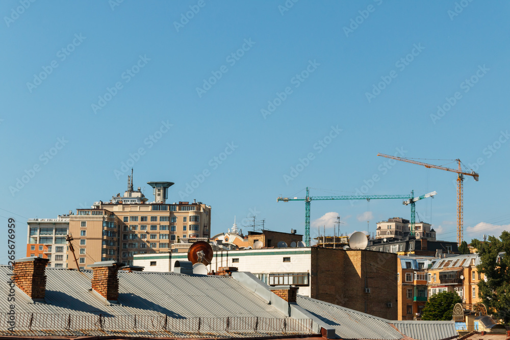 cityscape with roofs of houses and three construction cranes on the blue sky background with space for text. Turret slewing cranes, old and new architecture in Kyiv. cityscape with roofs of houses and three construction cranes on the blue sky background with space for text. Turret slewing cranes, old and new architecture in Kyiv.
