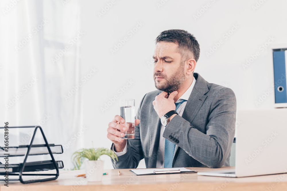 businessman sitting at desk, touching throat and holding glass of water in office