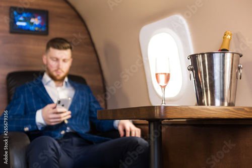 A young successful businessman in an expensive suit sits in the chair of a private jet with a glass of champagne in his hand and looks out the window photo