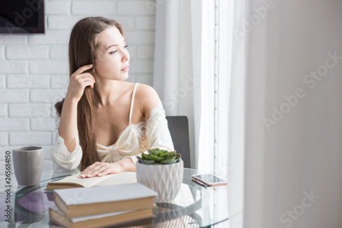 Beautiful young girl in a nightgown sits in the morning at the glass table, looks out the window and reads a book