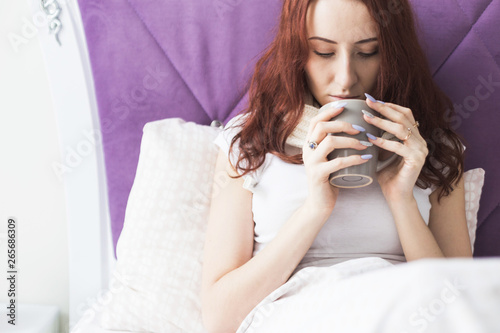 An attractive young brunette woman sits in her bed with a mug and pills. Colds and home treatment