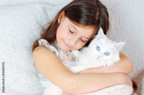 Home portrait of a little cute girl with a white cat Felis catus with blue eyes on a soft sofa