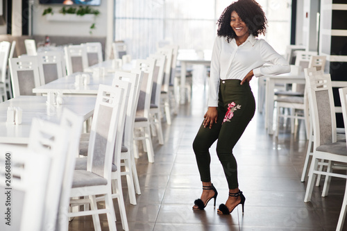 Сheerful business african american lady with afro hair, wear white blouse and green pants posed in cafe.