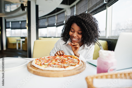   heerful business african american lady with afro hair  wear white blouse sitting at table  work with laptop in cafe  eat pizza and drink pink milkshake cocktail.