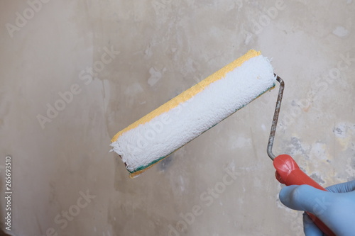 Male hand in blue glove holding a paint roller. Non-plastered concrete wall background. The concept of home repair, finishing works, preparation for priming the walls before painting.