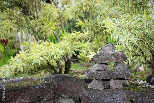 Small pile of seven rocks decoratively placed on a rock wall with a background of trees with long green and yellow leaves.