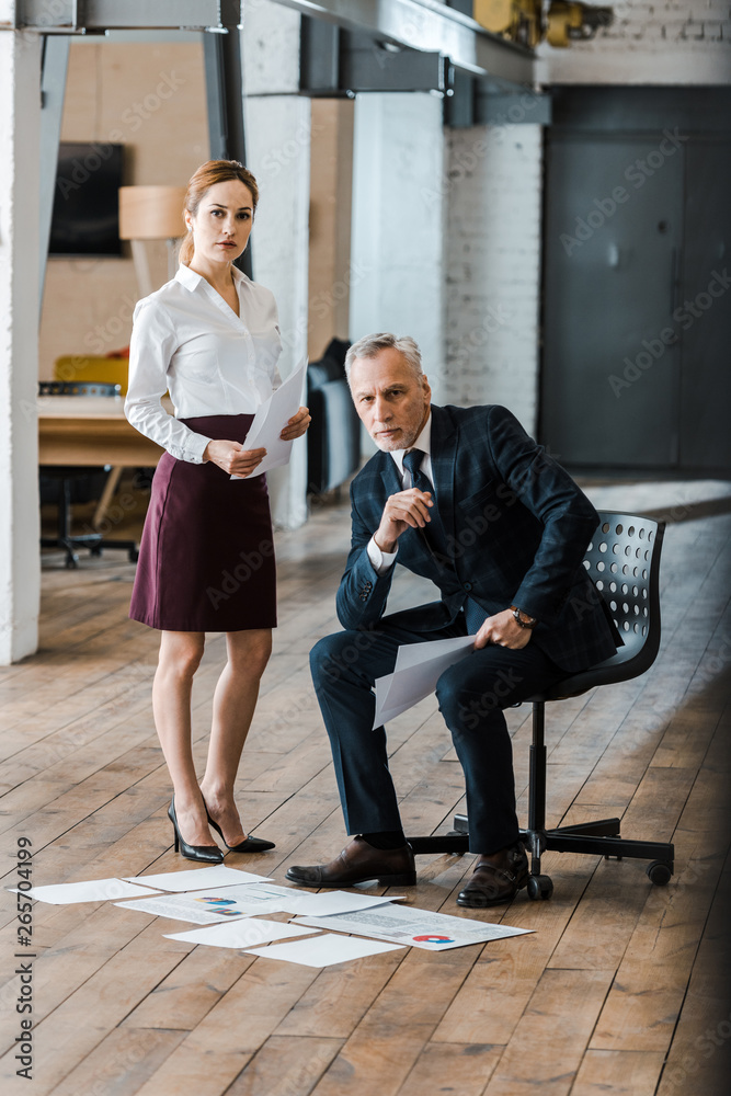 handsome man sitting on chair near businesswoman and charts and graphs on floor