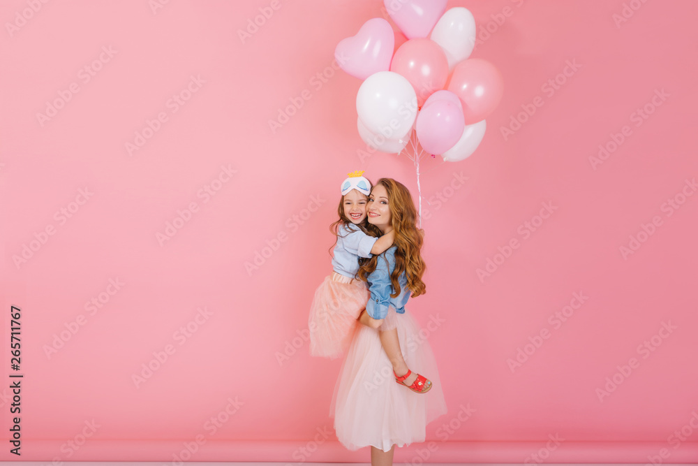 Portrait of attractive young woman holding cute little birthday girl with party balloons in her arms. Fascinating curly stylish girl in lush skirt hugging her sister with love on pink background