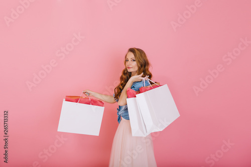 Attractive curly girl in denim shirt holding big white bags from clothing store isolated on pink background. Charming long-haired young woman in cute outfit posing with packages after shopping.