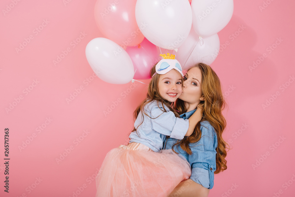 Portrait of adorable long-haired birthday girl in sleep mask holding colorful balloons and embracing her stylish mom after event. Charming mother posing with pretty daughter at party in pink room Portrait of adorable long-haired birthday girl in sleep mask holding colorful balloons and embracing her stylish mom after event. Charming mother posing with pretty daughter at party in pink room