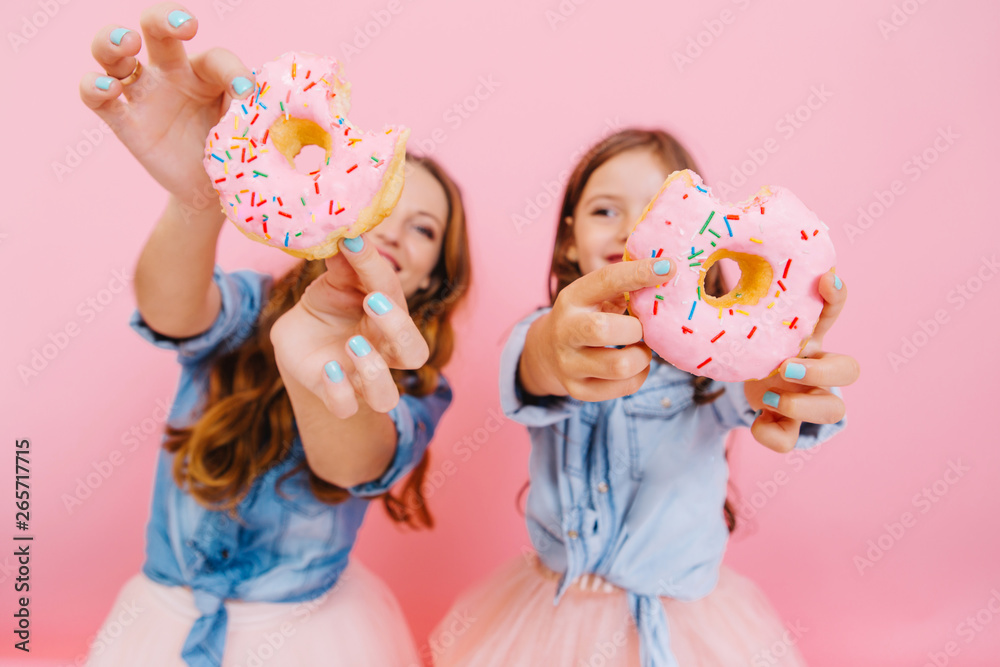 Young cheerful mother and cute smiling daughter having fun with tasty donuts waiting for tea party with family. Little girl with her mom showing doughnuts wich they cooked together and laughing - obrazy, fototapety, plakaty 