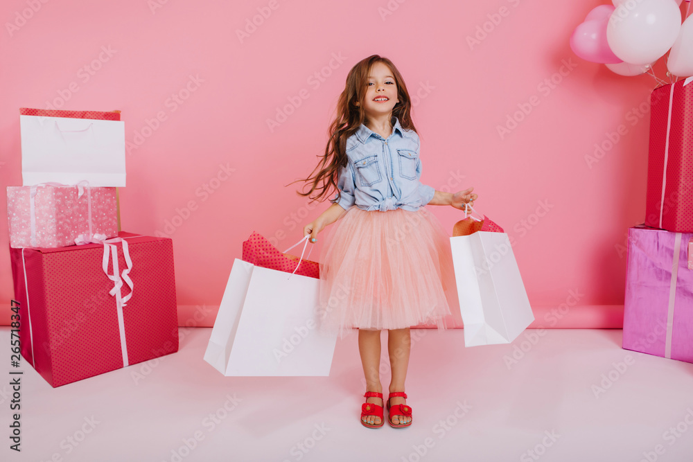 Pretty joyful little girl in tulle skirt  walking with packages suround giftboxes on pink background. Lovely sweet moments of cute child celebrating birthday, expressing positivity to camera