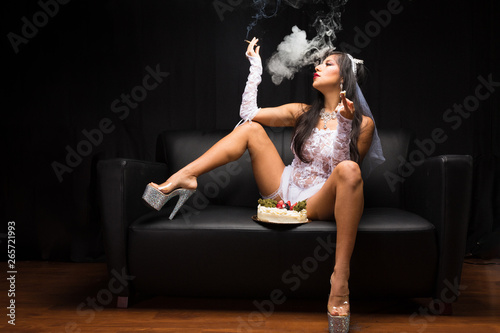 Hispanic woman in bridal lingerie smokes and poses with cannabis on a cake under soft overhead light