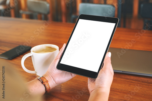 Mockup image of hands holding black tablet pc with blank white screen with coffee cup on wooden table