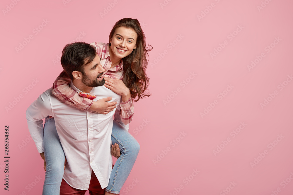 Photo of playful man gives piggyback ride to girlfriend, have fun together, smile happily, pose over pink background with blank space for your text information. Happy man carries woman on back Photo of playful man gives piggyback ride to girlfriend, have fun together, smile happily, pose over pink background with blank space for your text information. Happy man carries woman on back