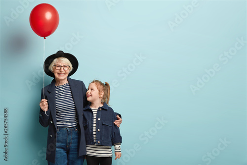 Indoor shot of fashionable senior woman embraces little child, enjoy spending time together, celebrate first day at school, hold air balloon, pose over blue background with blank space on right photo