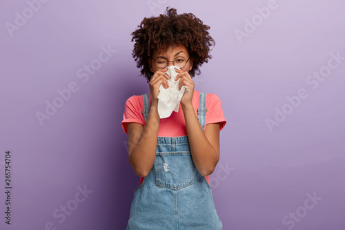 Obraz na plátne Portrait of sick African American woman sneezes in white tissue, suffers from rhinitis and running nose, has allergy on something, looks unhealthy, feels unwell