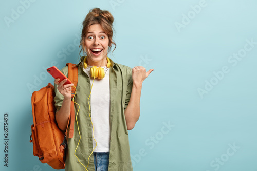 Joyful European girl checks nootification or email box on cellular, enjoys listening favourite music in headphones, has surprised happy reaction on something aside, points thumb at free space photo