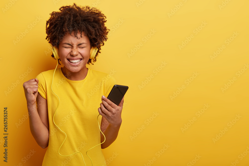 Charming satisfied dark skinned female holds smartphone, clenches fist, closes eyes from pleasure, enjoys mini concert in ears, wears yellow t shirt, stands over studio background with blank space Charming satisfied dark skinned female holds smartphone, clenches fist, closes eyes from pleasure, enjoys mini concert in ears, wears yellow t shirt, stands over studio background with blank space