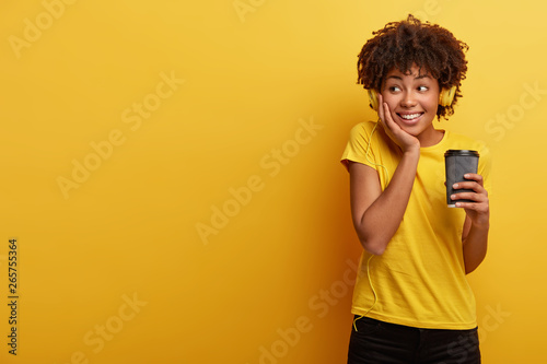 Time for music. Joyful dark skinned female holds coffee to go in disposable cup, listens pleasant melody in headphones, looks happily away, wears casual yellow t shirt, poses in studio, blank space