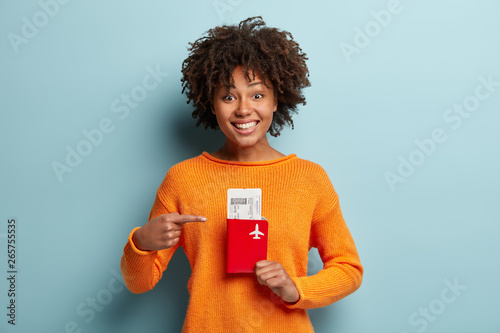 Photo of glad dark skinned curly female traveller points at tickets and passport, rejoices having trip abroad on summer holidays, prepares for departure, flies by plane first time. Voyage, travelling