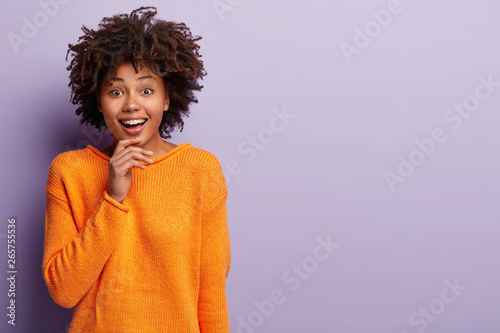 Beautiful woman with Afro hairstyle smiles gently at camera  touches chin  looks happily at camera  wears orange jumper  isolated over purple background with copy space aside for your promotion