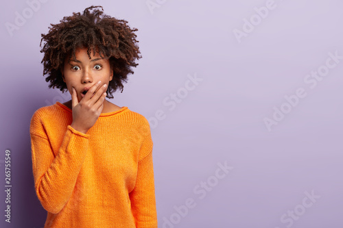 People and emotions concept. Shocked terrified woman covers widely opened mouth  hears terrible rumor  gazes with fright  wears orange jumper  models over purple background with blank space.