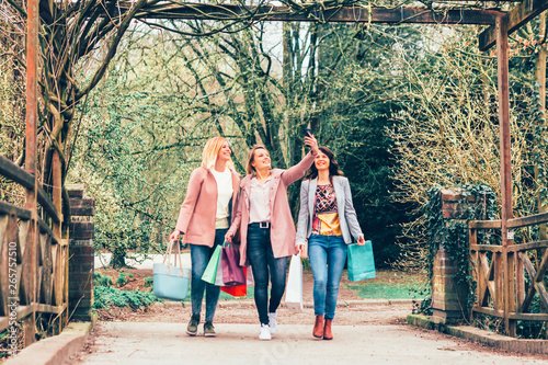 three young women in a park smiling and caring shopping bags they take a selfie while on walking - togetherness, free time, friendship and consumption concept © Carlo