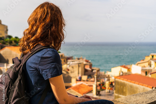 Female tourist with a backpack sitting on the city walls, relaxing and enjoying the view over the colorful old town of Riomaggiore, in Cinque Terre, Italy, a famous tourist attraction.