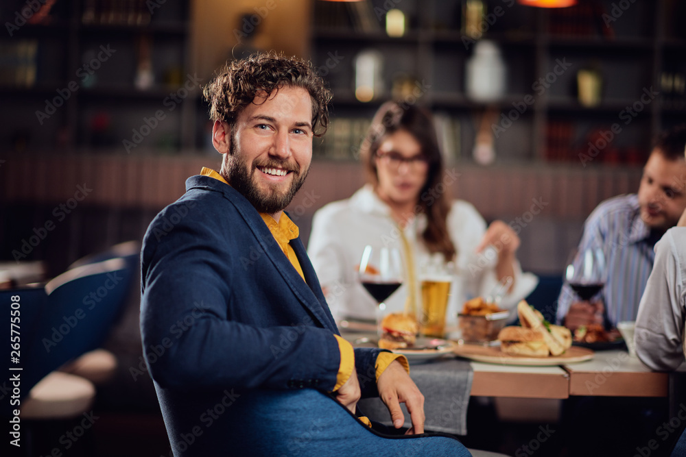 Smiling attractive Caucasian man sitting in restaurant and looking over shoulder. In background his friends having dinner.