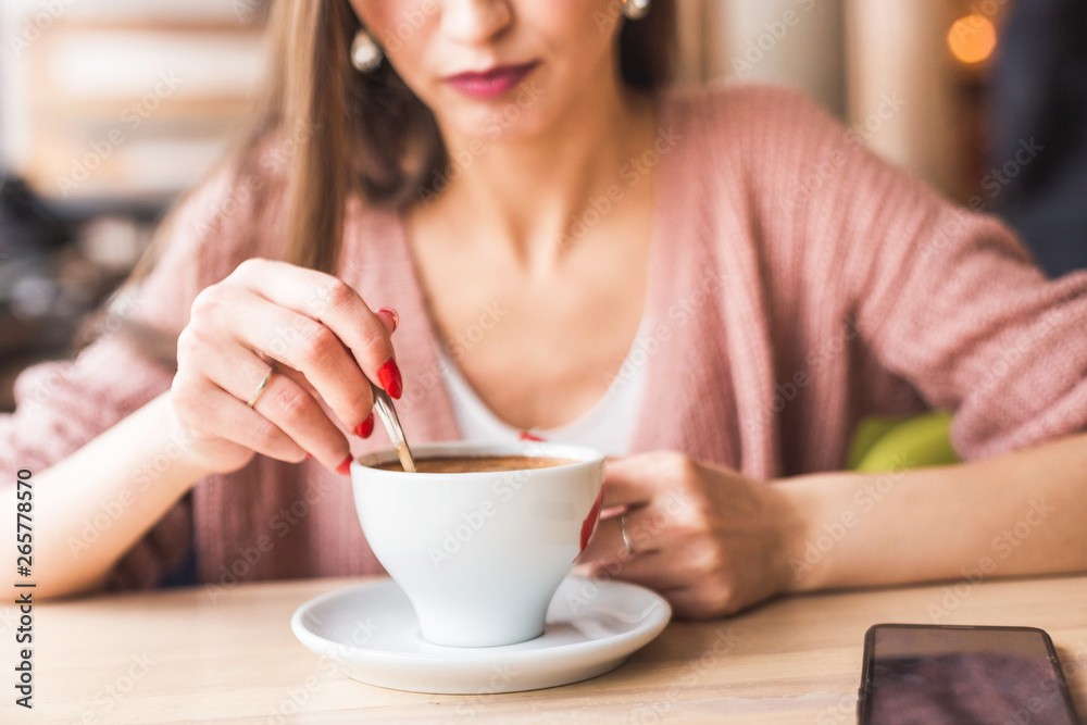 Attractive young woman sits at a table in a cafe with a cup of coffee and mixes it with a spoon Attractive young woman sits at a table in a cafe with a cup of coffee and mixes it with a spoon