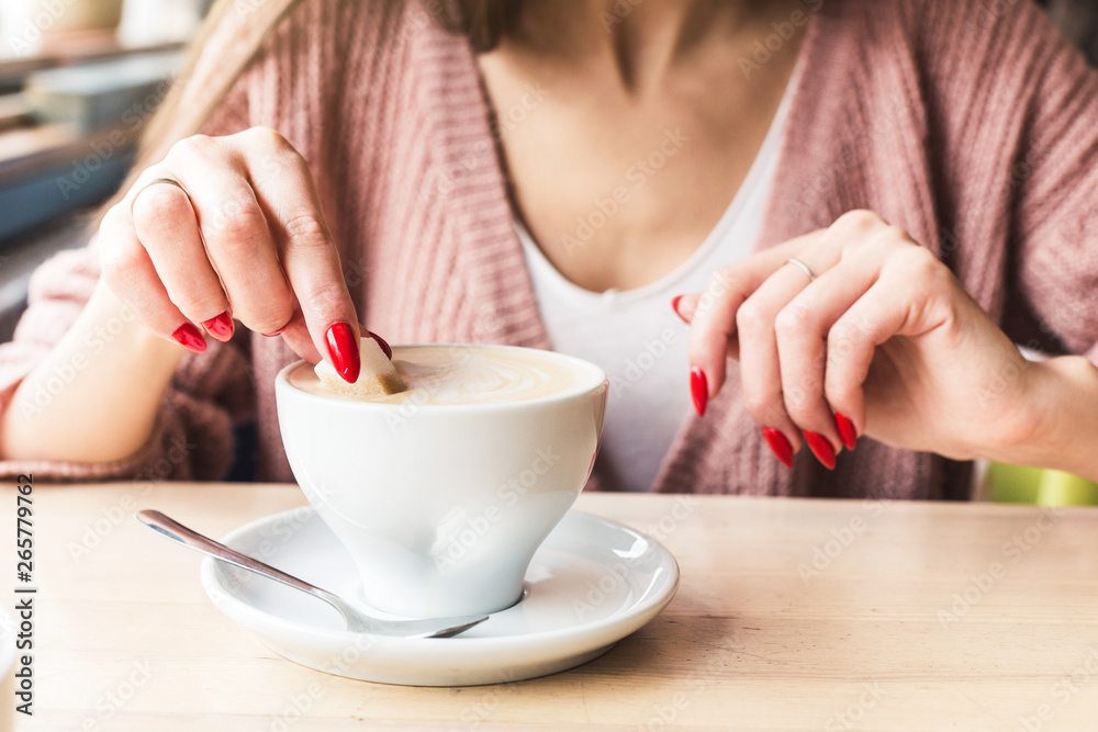A girl dunks a piece of sugar in coffee. Closeup of hands with red nails make-up and cappuccino cup