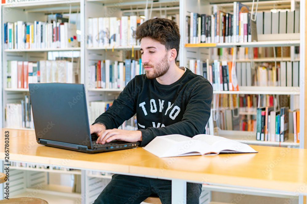 Young student sitting in front of the computer with books at the table concentrating writing in the library Young student sitting in front of the computer with books at the table concentrating writing in the library