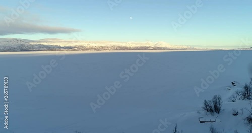 Boreal lake, C4k aerial, rising, drone shot, over branches and arctic, icy Tornetrask, towardsmountains, near Abisko park, at golden hour, on a sunny winter evening, Lapland, Norrbotten, Sweden