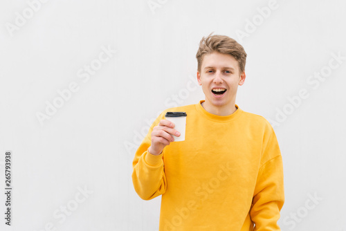 Joyful guy with a cup of coffee on the background of a white wall, looking into the camera. An expressive young man holds a paper cup in his hands, isolated against a background of a light wall.