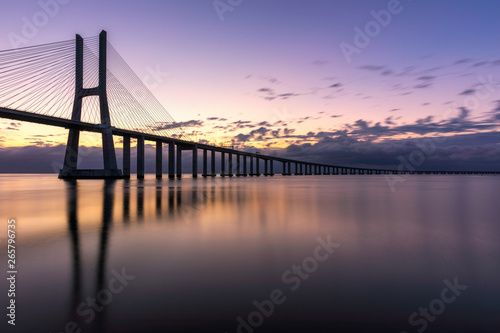 Vasco da Gama bridge at sunrise in Lisbon, Portugal. Vasco da Gama Bridge is a cable-stayed bridge flanked by viaducts and rangeviews that spans the Tagus River in Parque das Nações in Lisbon.