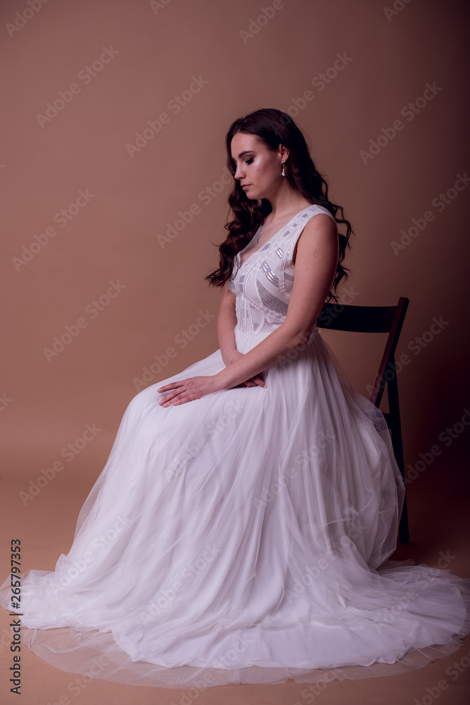 Fototapeta premium Beautiful young girl in a flying white dress. Flowing fabric. bride in white dress on beige background. gorgeous brunette on a studio background. Isolate