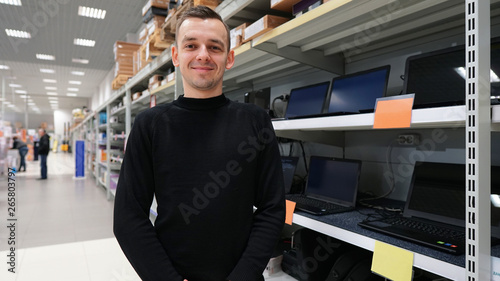 Portrait of best a Professional Expert Consultant Smiles and Looks into Camera as Stands in the Bright, Modern Electronics Store Full of Latest Models of TV Sets, Cameras, Tablets and other Devices. 