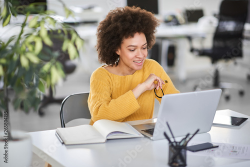 Smiling mixed race businesswoman holding eyeglasses and looking at laptop while sitting in modern office.