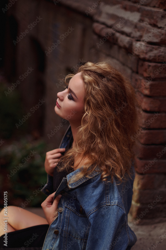 Obraz premium Stylish handsome curly blonde wearing chic little black dress and jeans jacket. Glamour girl posing and smiling at camera.
