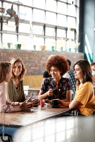 Four Young Female Friends Meeting Sit At Table In Coffee Shop And Talk