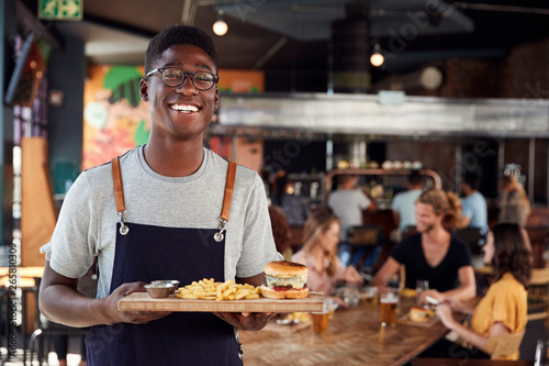 Portrait Of Waiter Serving Food To Customers In Busy Bar Restaurant photo