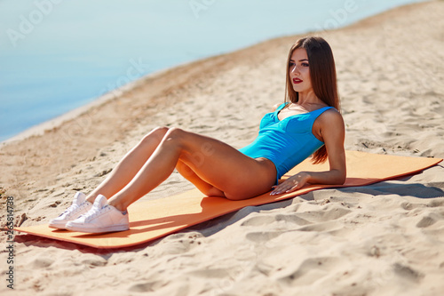 Portrait of beautiful sporty woman in swimwear doing workout fitness stretching exercises on the sand beach at early morning. Female sporty muscular body. Bright sunny light.