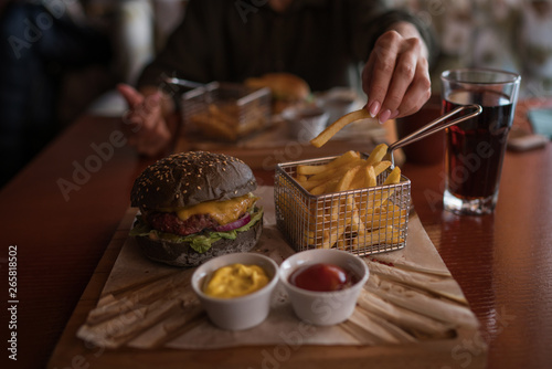 Tasty burger and sauce on wooden tray. Woman eating burger and chips in cafe. People and eating concept. Hamburger  french fries  ketchup  mustard on a wooden board. Toned image.
