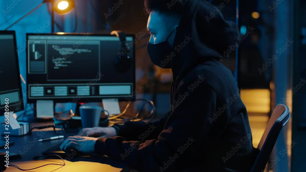 cybercrime, hacking and technology concept - asian male hacker in mask writing code or using computer virus program for cyber attack in dark room at night cybercrime, hacking and technology concept - asian male hacker in mask writing code or using computer virus program for cyber attack in dark room at night