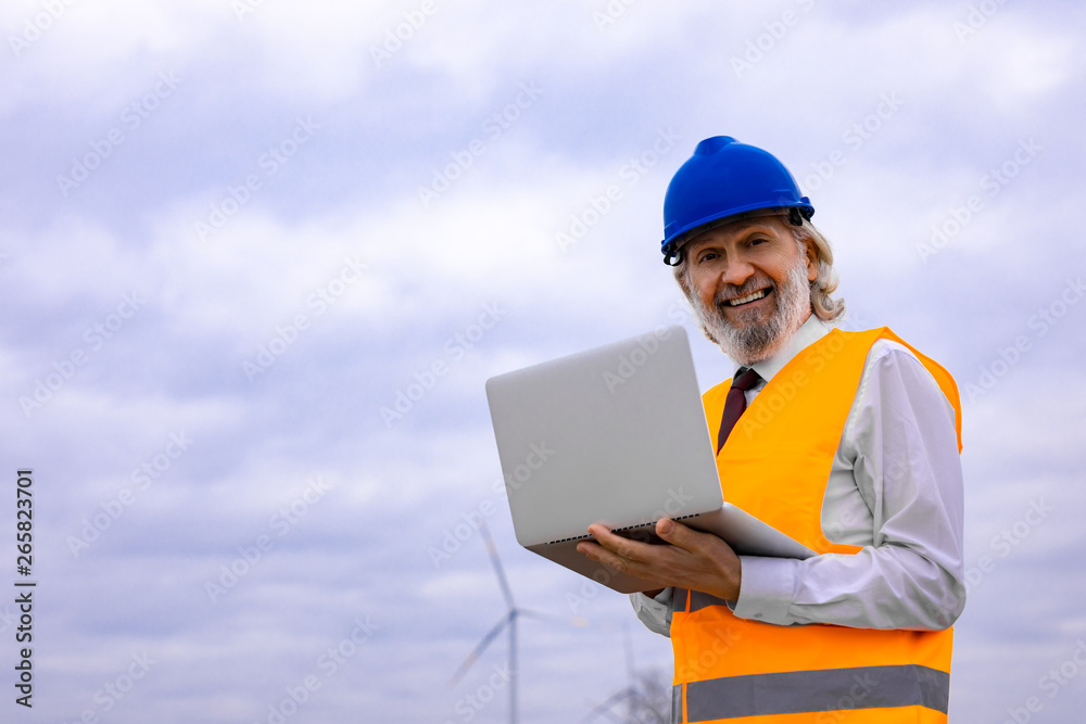 Senior wind turbine engineer with laptop in safety gear on the field