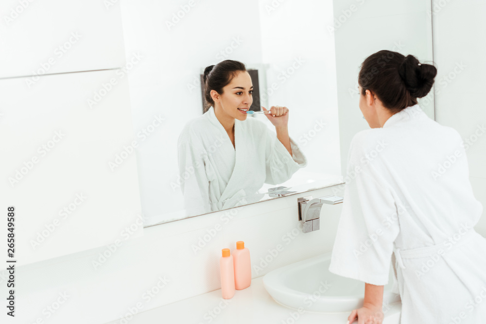 selective focus of cheerful woman looking at mirror while brushing teeth in bathroom