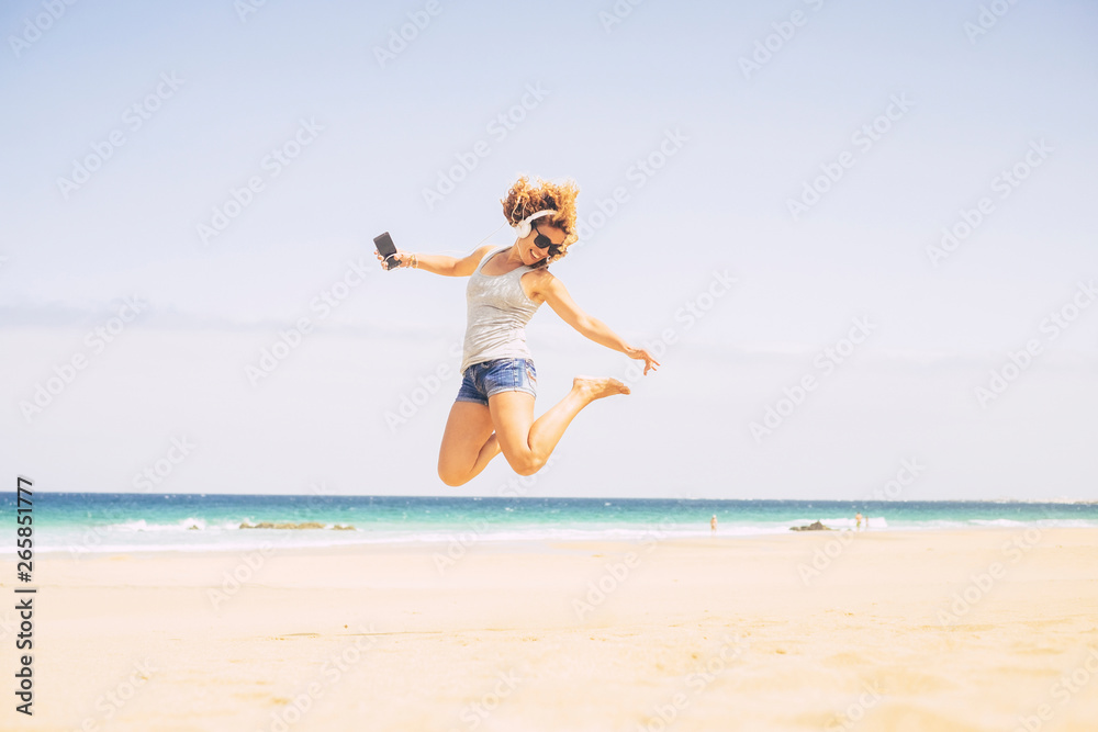 People happines concept for summer holiday vacation leisure activity at the beach - young crazy beautiful blonde woman jump with phone and headphones with sea in background People happines concept for summer holiday vacation leisure activity at the beach - young crazy beautiful blonde woman jump with phone and headphones with sea in background