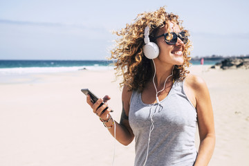 Cheerful beautiful young people blonde woman curly hair enjoying the summer holiday vacation at the beach listineg music from modern phone and headphones - sea and sand in background