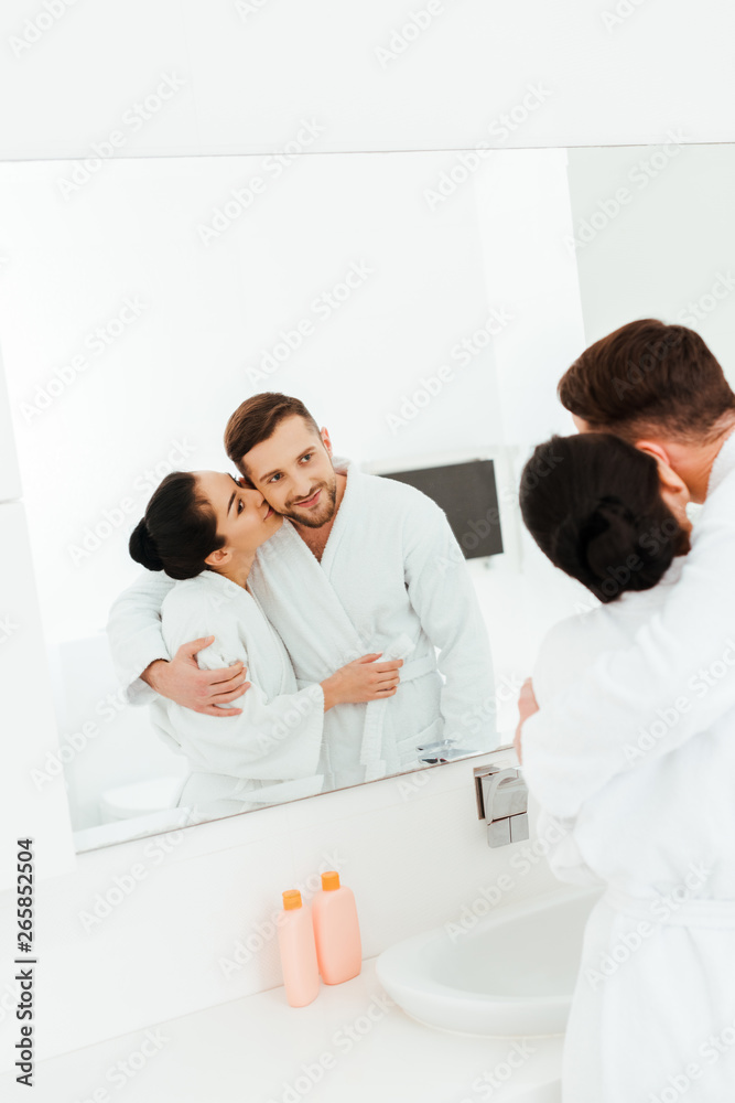 selective focus of happy woman hugging and kissing cheek of man while looking at mirror selective focus of happy woman hugging and kissing cheek of man while looking at mirror